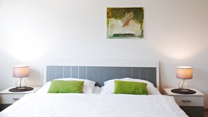 Een netjes opgemaakt bed met wit beddengoed en groene kussens, met aan weerszijden twee nachtkastjes met lampen, tegen een witte muur met een schilderij erboven.