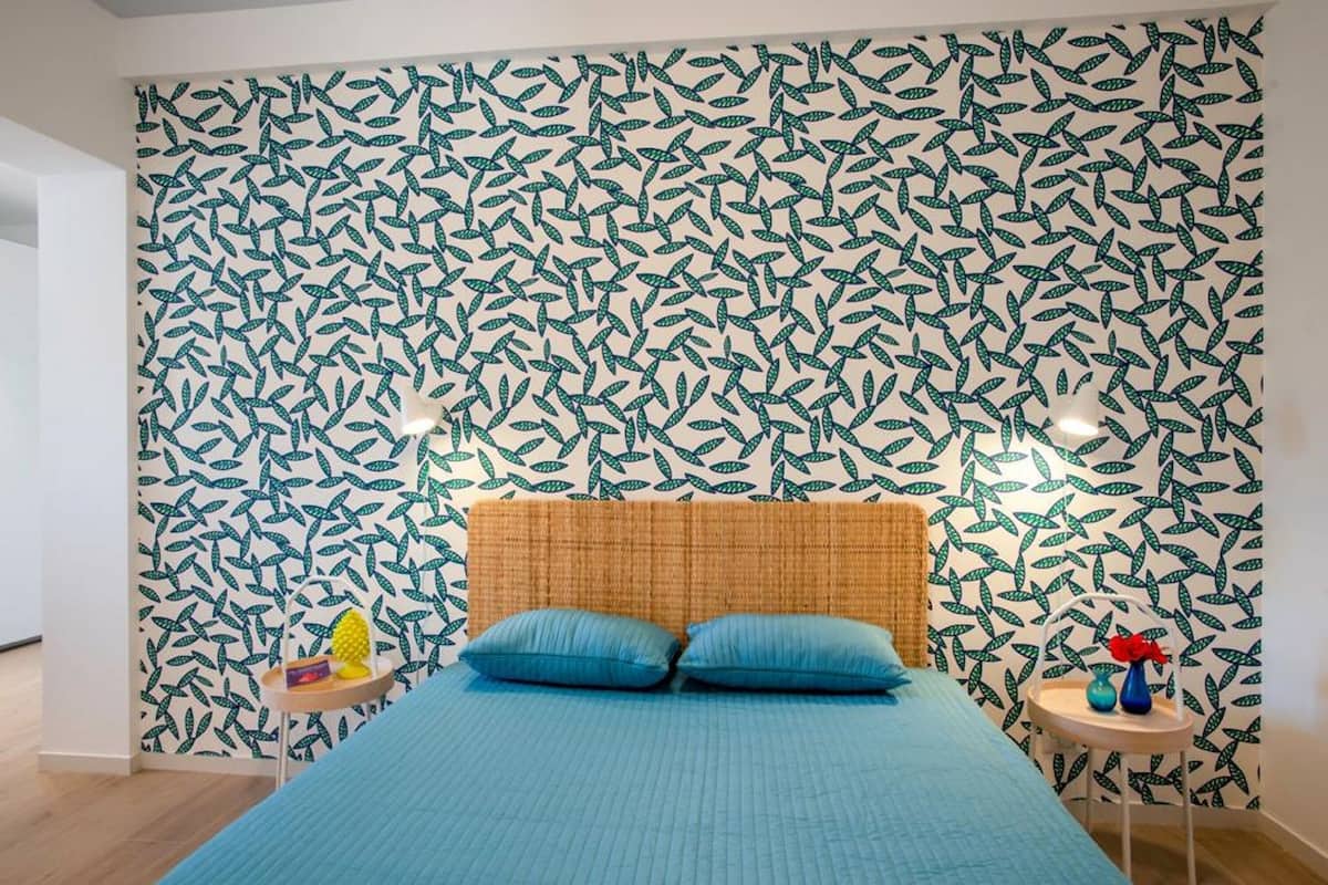 Ein Schlafzimmer mit einem Bett, blauer Bettwäsche, Tapete mit Blättermotiv, einem gewebten Kopfteil, Nachttischen mit Dekoration und einem kleinen Beistelltisch mit einer Ananas und einer Vase.