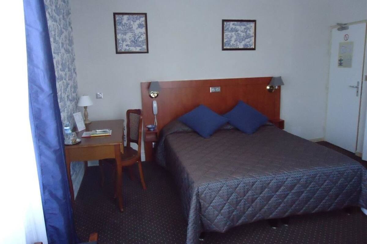 A hotel room with a bed, a desk, two chairs, a door, and two framed pictures on the wall.