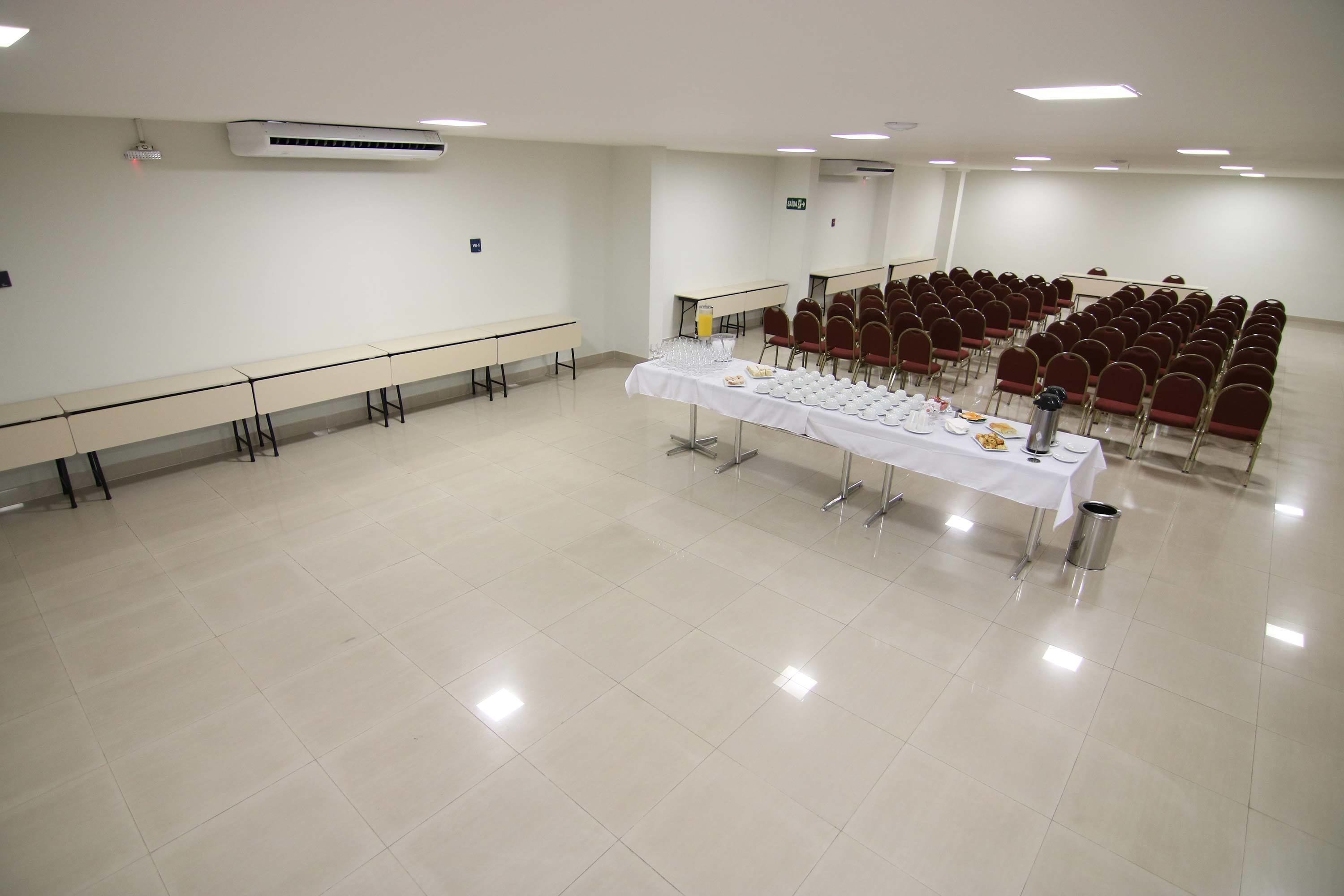 Un salón de conferencias con una larga mesa dispuesta para una comida, hileras de sillas rojas y un equipo de aire acondicionado en la pared.