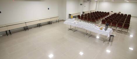 Un salón de conferencias con una larga mesa dispuesta para una comida, hileras de sillas rojas y un equipo de aire acondicionado en la pared.