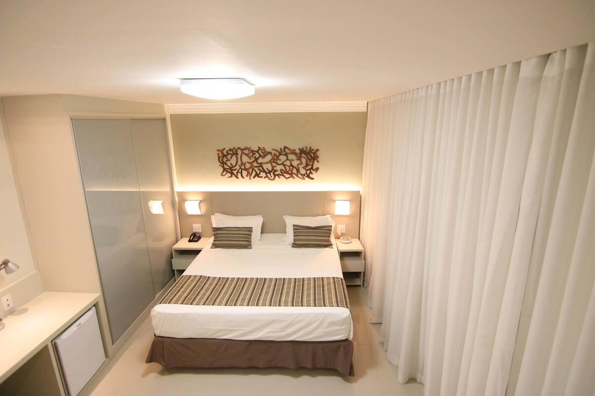 Ein Hotelzimmer mit Bett, Nachttischen, Schreibtisch, Spiegel und Vorhang.
