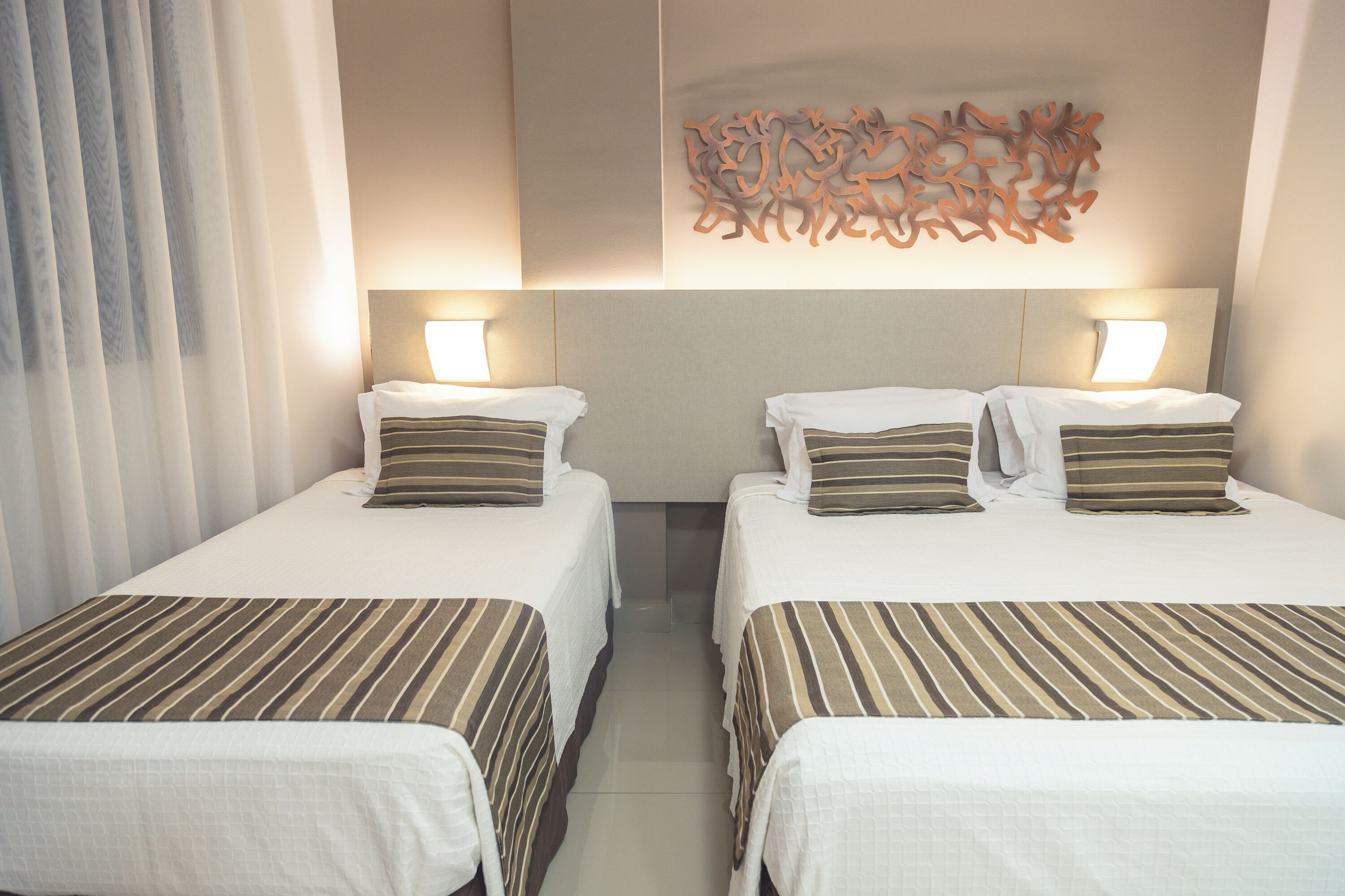 Una habitación de hotel con dos camas individuales, cada una con colcha y almohadas a rayas, un cuadro abstracto en la pared y una lámpara de noche.