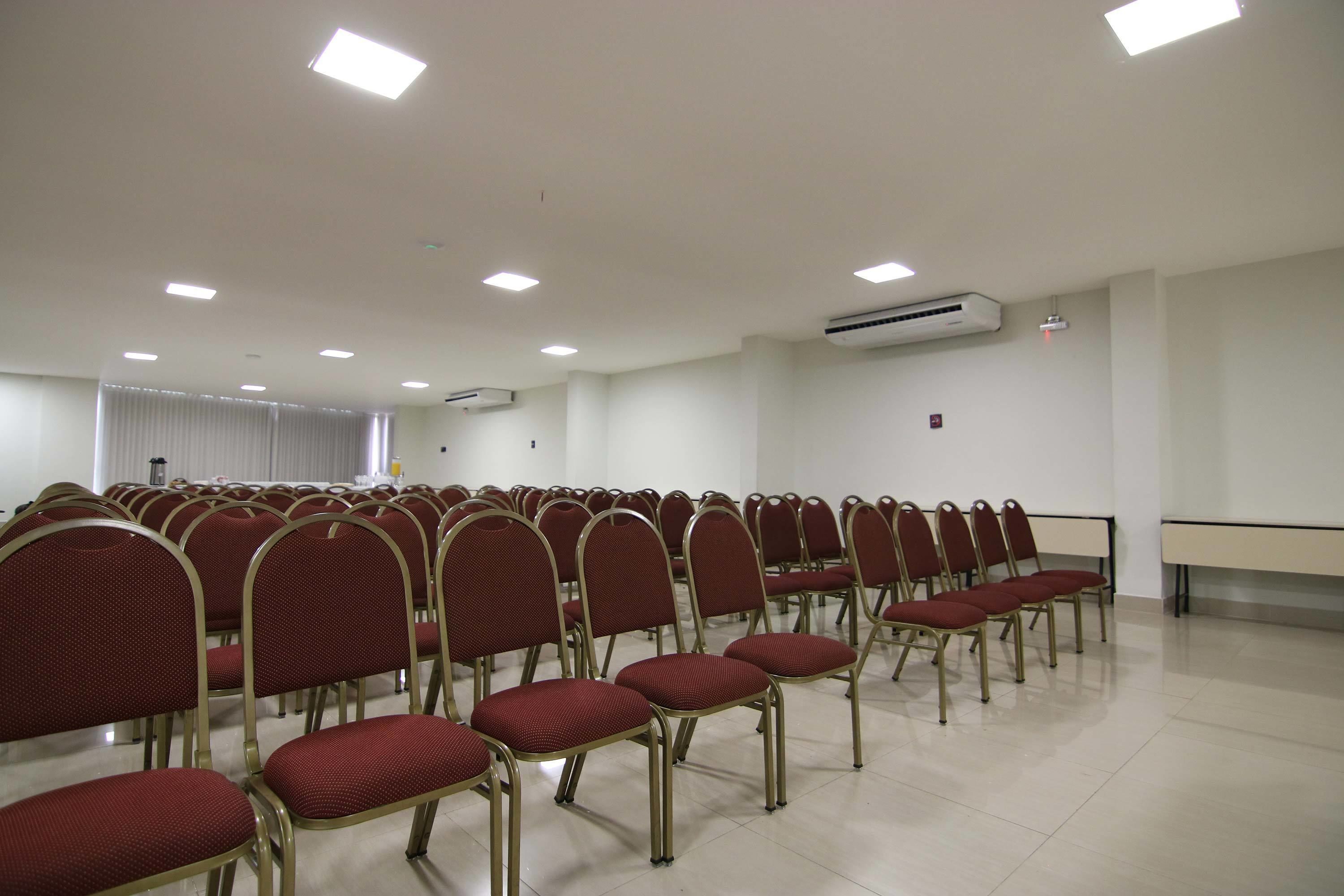Un salón de conferencias con filas de sillas rojas, paredes blancas e iluminación en el techo.