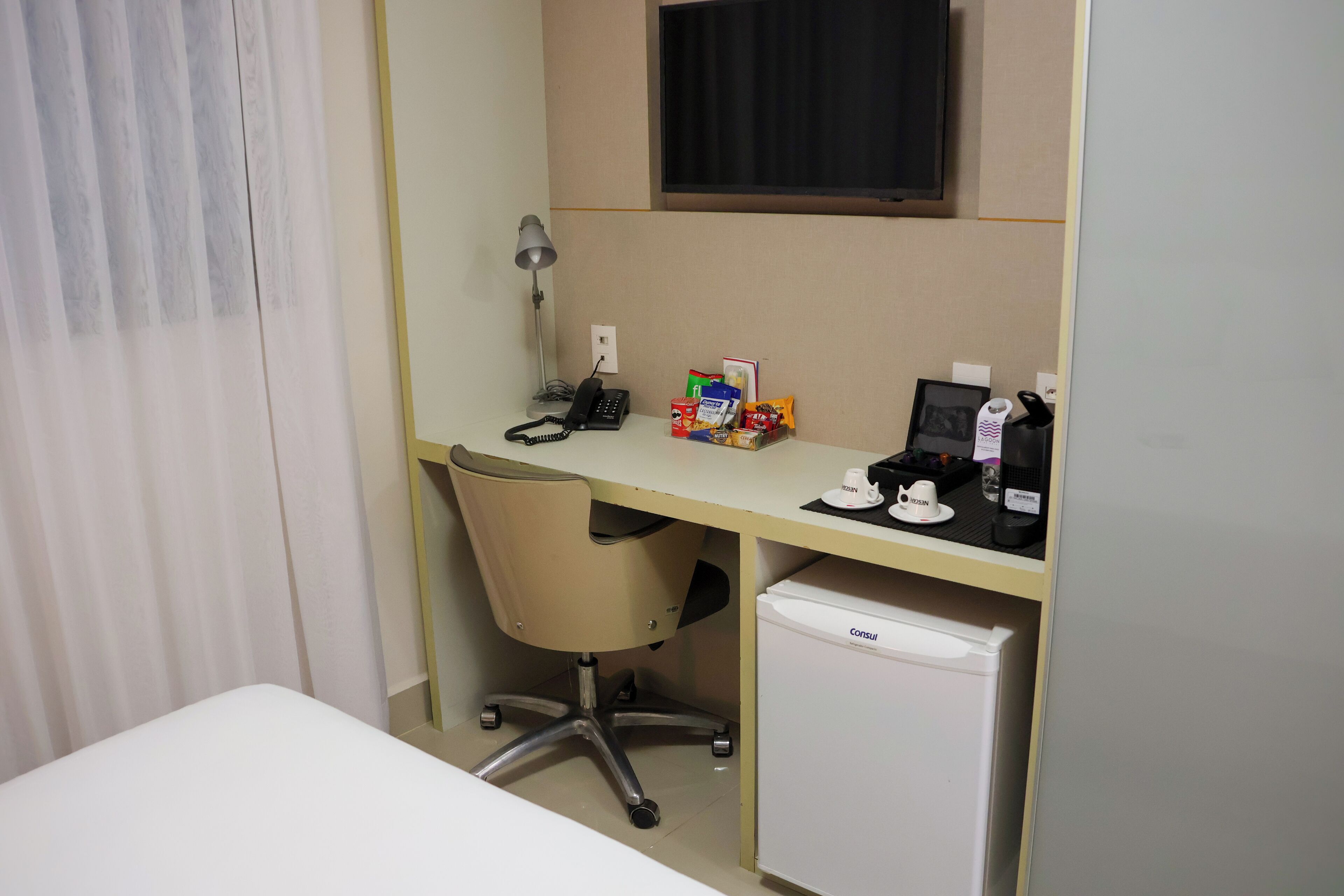 A hotel room with a desk, chair, and a small refrigerator.
