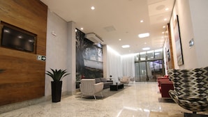 A modern lobby with a wooden accent wall, a flat-screen TV, seating area with patterned chairs, a sofa, and a large window with curtains. - Lagoon Prime Hotel (Lagoa Santa)