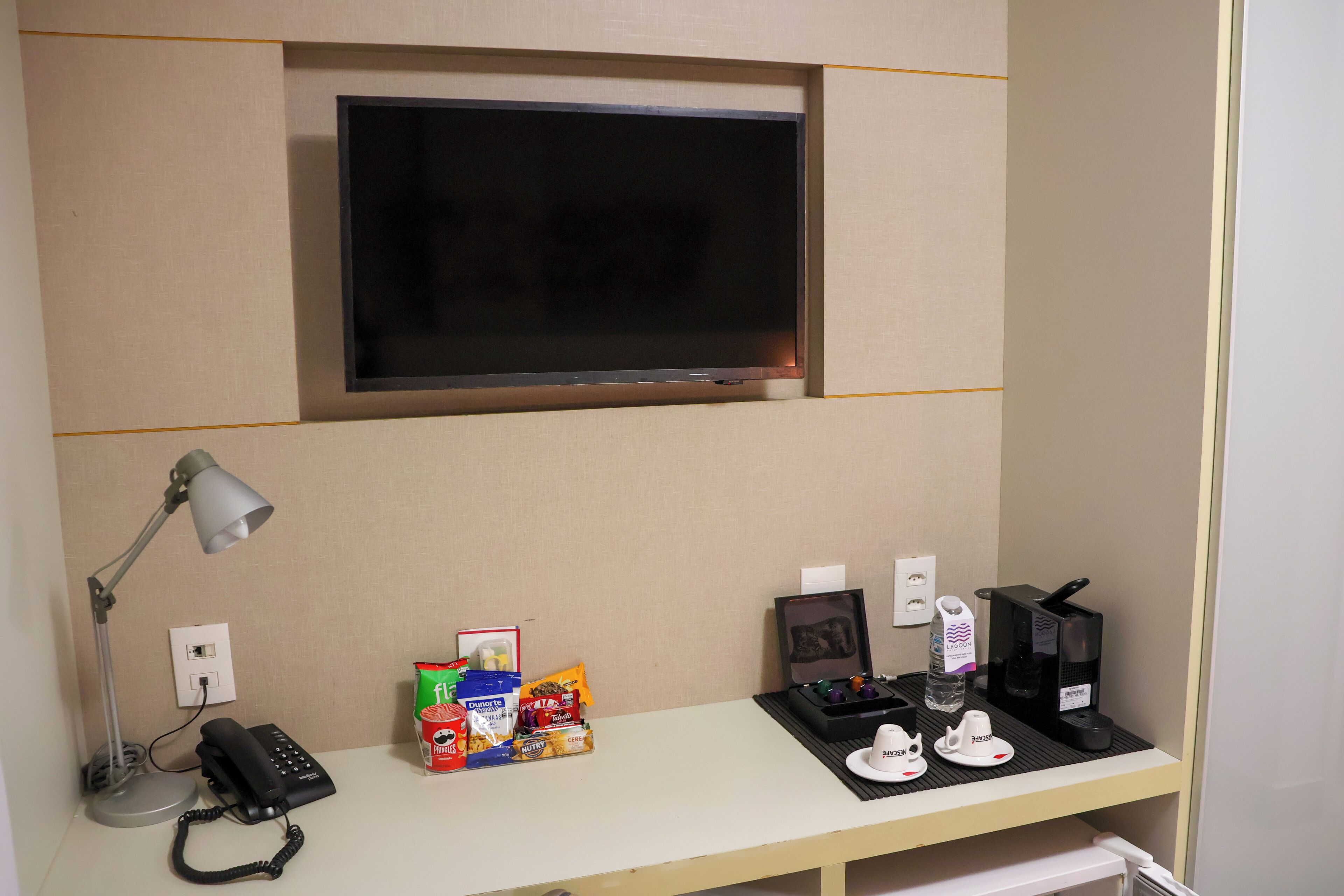 Quarto de hotel com escrivaninha, televisão na parede, luminária de mesa, telefone, lanches, cafeteira e um pequeno porta-retrato.