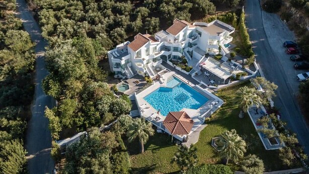 An aerial view of a modern resort with multiple buildings, a central swimming pool, and lush greenery surrounding the property. - Villa Life (Chania)