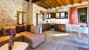 A cozy living room with a sofa, a coffee table, and a decorative mirror. Adjacent is a kitchen with wooden cabinets, a refrigerator, and a dining area with a table and chairs. - Villa Life (Chania)