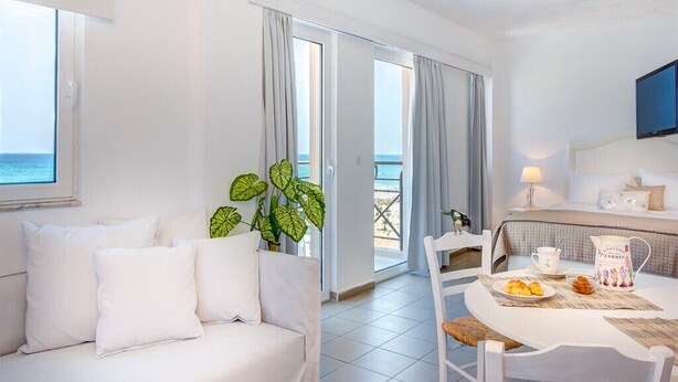 A hotel room with a white sofa, a dining table with a pitcher and plate of pastries, a TV mounted on the wall, and a view of the sea through the window. - Aqua Marine (Kissamos)