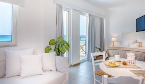 A hotel room with a white sofa, a dining table with a pitcher and plate of pastries, a TV mounted on the wall, and a view of the sea through the window. - Aqua Marine (Kissamos)