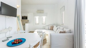 A modern, white-themed living room with a sofa, a dining table with a blue plate of fruit, and a TV mounted on the wall. - Aqua Marine (Kissamos)