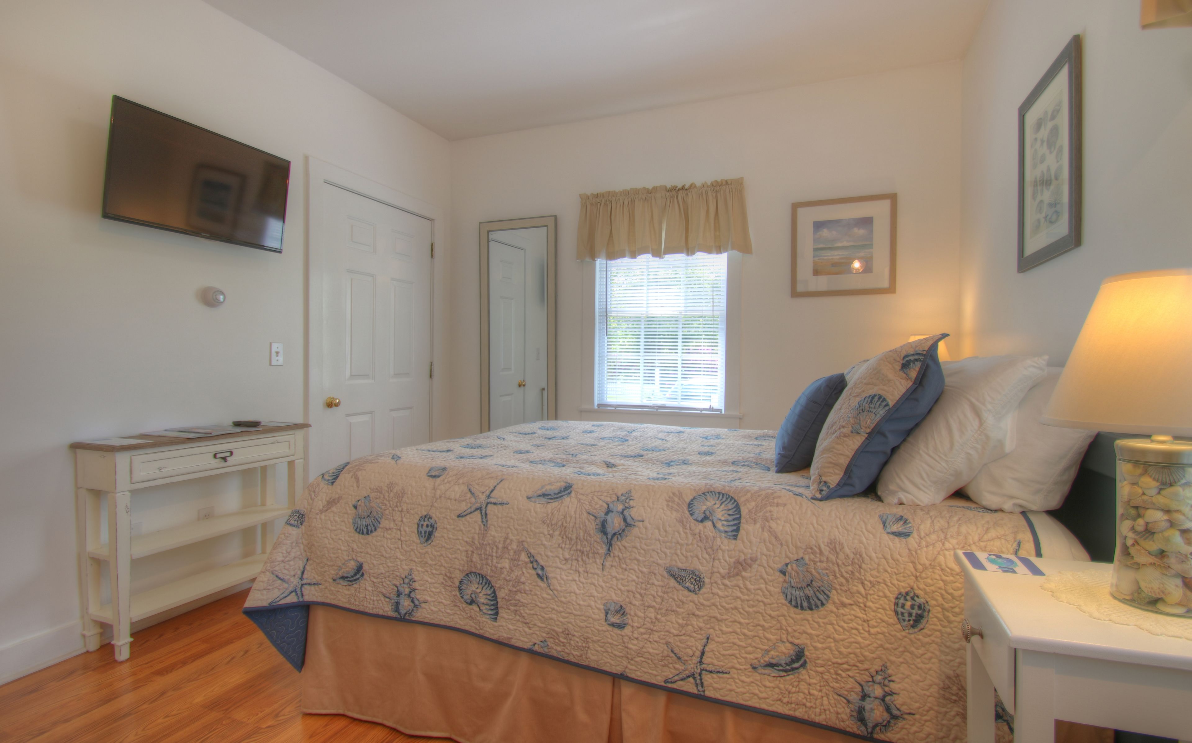 A bedroom with a bed, a desk, a window with blinds, and a wall-mounted TV.
