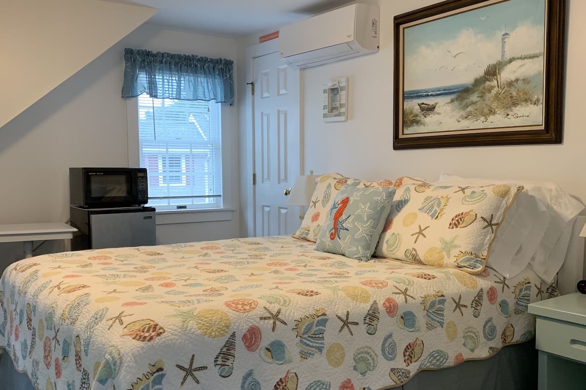 Ein Schlafzimmer mit Bett, Nachttisch, Fenster mit Vorhängen, einem Gemälde und einer Deckenleuchte.