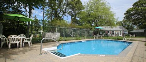 Una piscina rectangular con una escalera metálica y un trampolín, rodeada por una valla. Hay sillas y una mesa exteriores, con un paraguas verde.