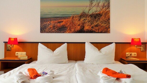 A hotel room with a large bed, two pillows, a nightstand with a lamp, and a painting of a beach scene on the wall. - Hotel Seeblick (Klausdorf)