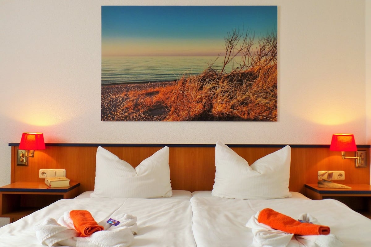 A hotel room with a large bed, two pillows, a nightstand with a lamp, and a painting of a beach scene on the wall.