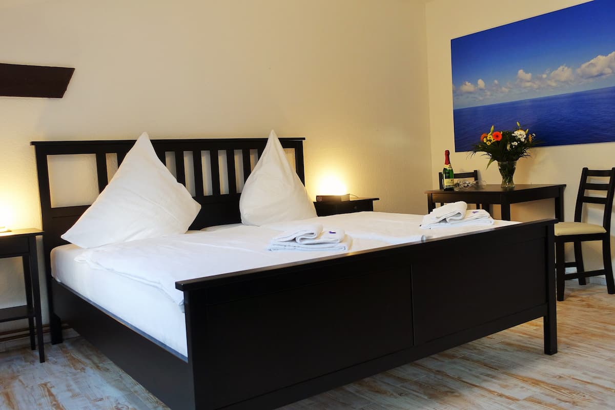 A neatly made bed with white linens and pillows, a bedside table with a bottle and glasses, and a painting of the sea on the wall.