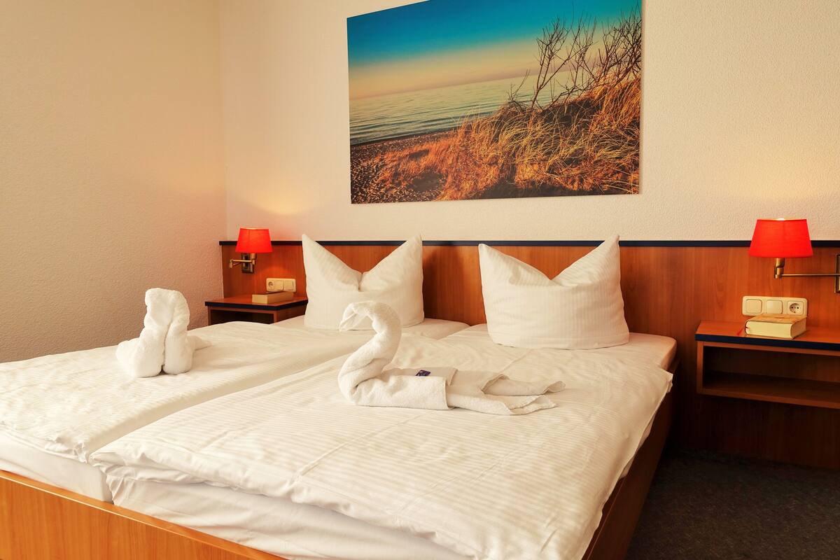A double bed with white linens and pillows, two bedside tables with lamps, a wall-mounted painting, and a red lamp on the nightstand.