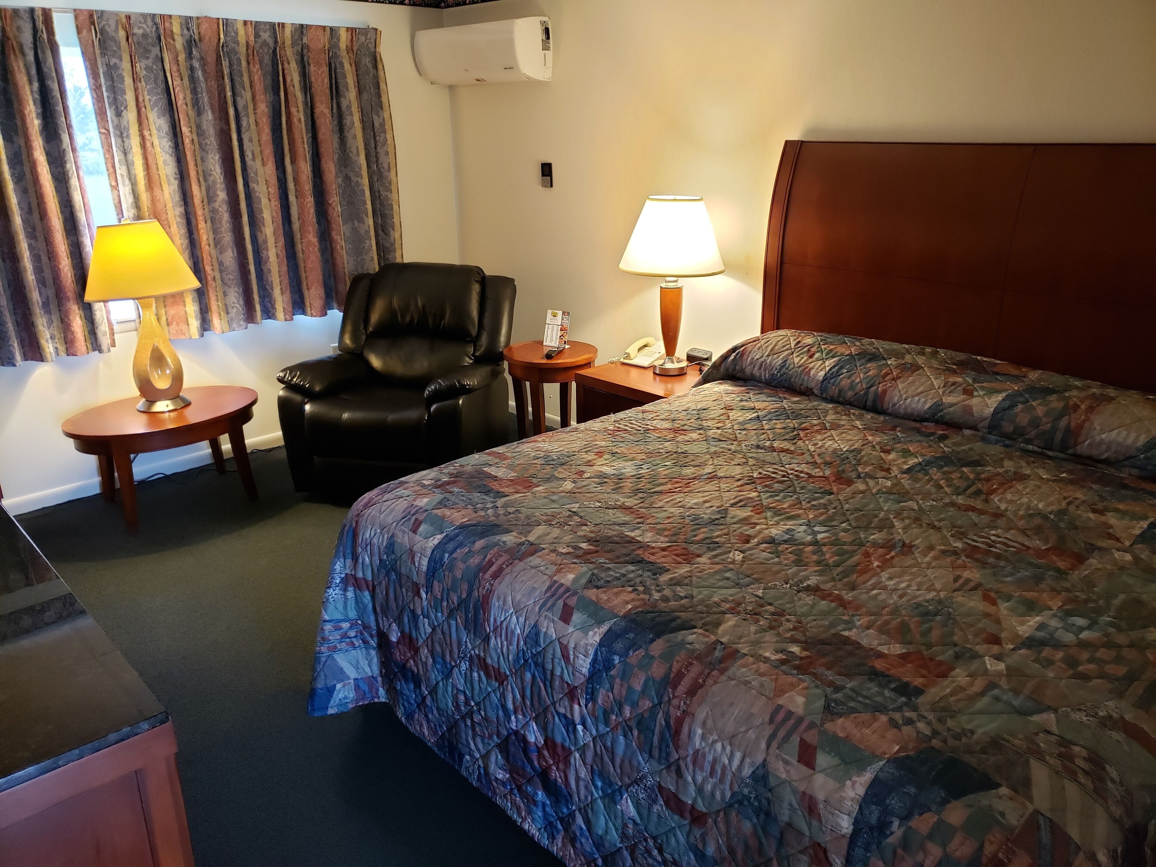 A hotel room with a large bed, a leather chair, two bedside tables with lamps, a telephone, and an air conditioning unit on the wall.