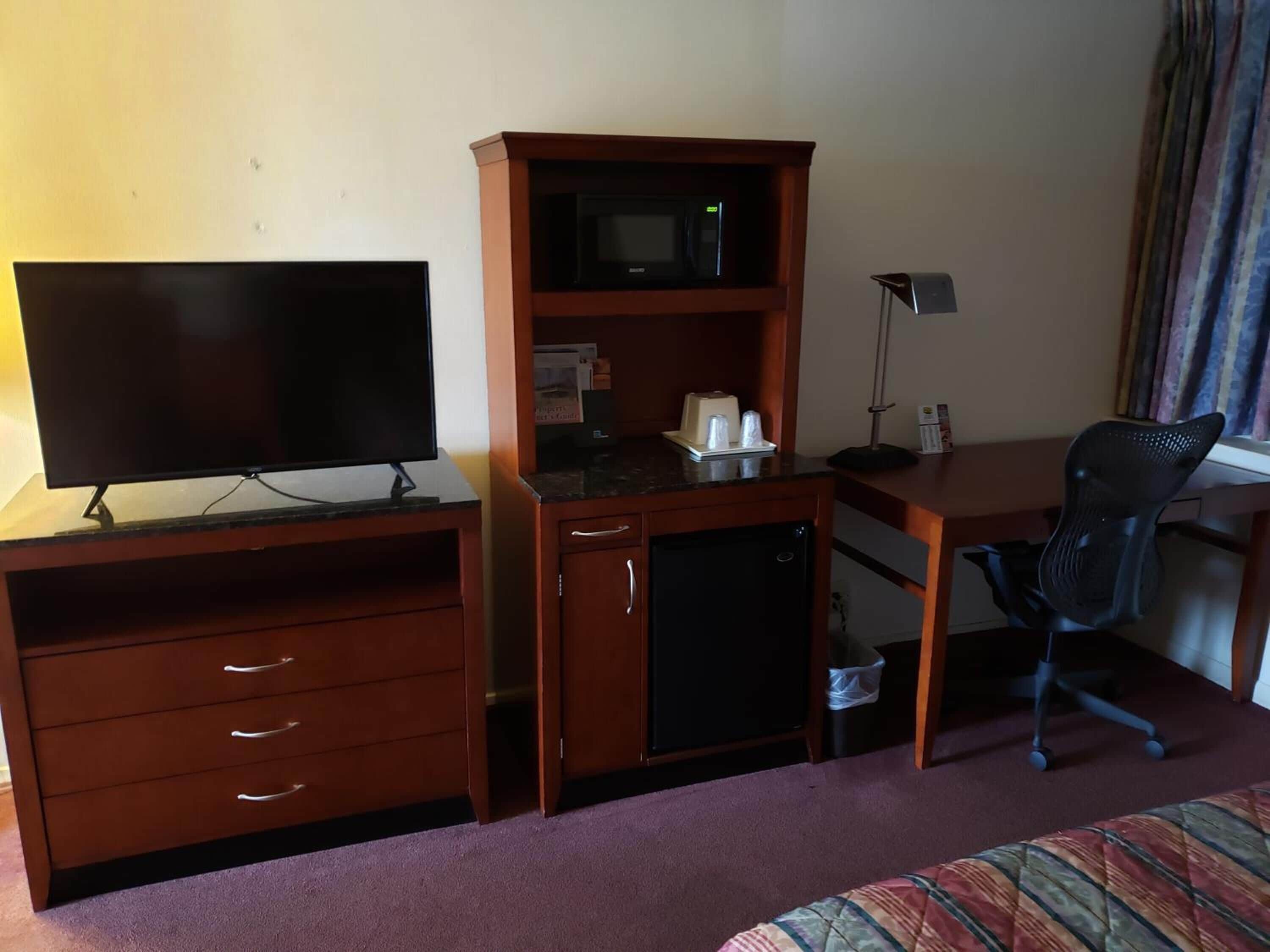 A hotel room with a wooden desk, a television, a microwave, a refrigerator, a chair, and a bed.
