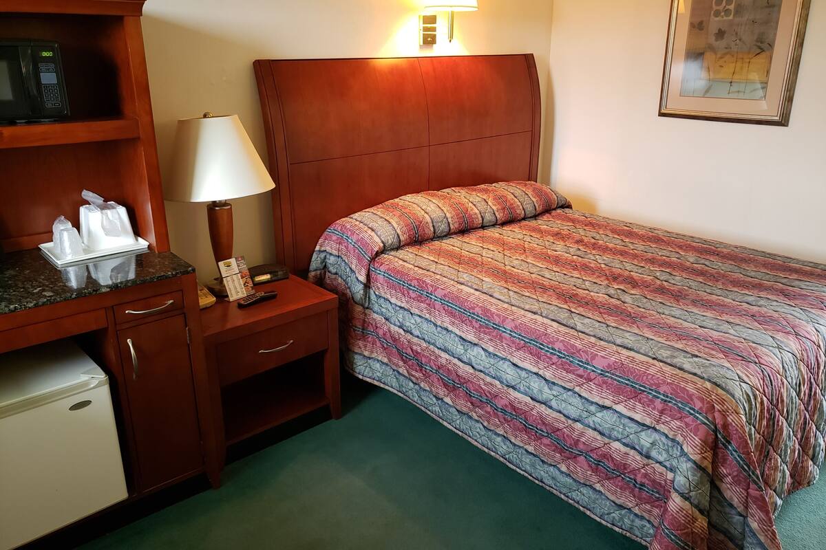 A hotel room with a wooden headboard, a bedside table with a lamp, a microwave, a mini fridge, and a bed with a colorful quilt.