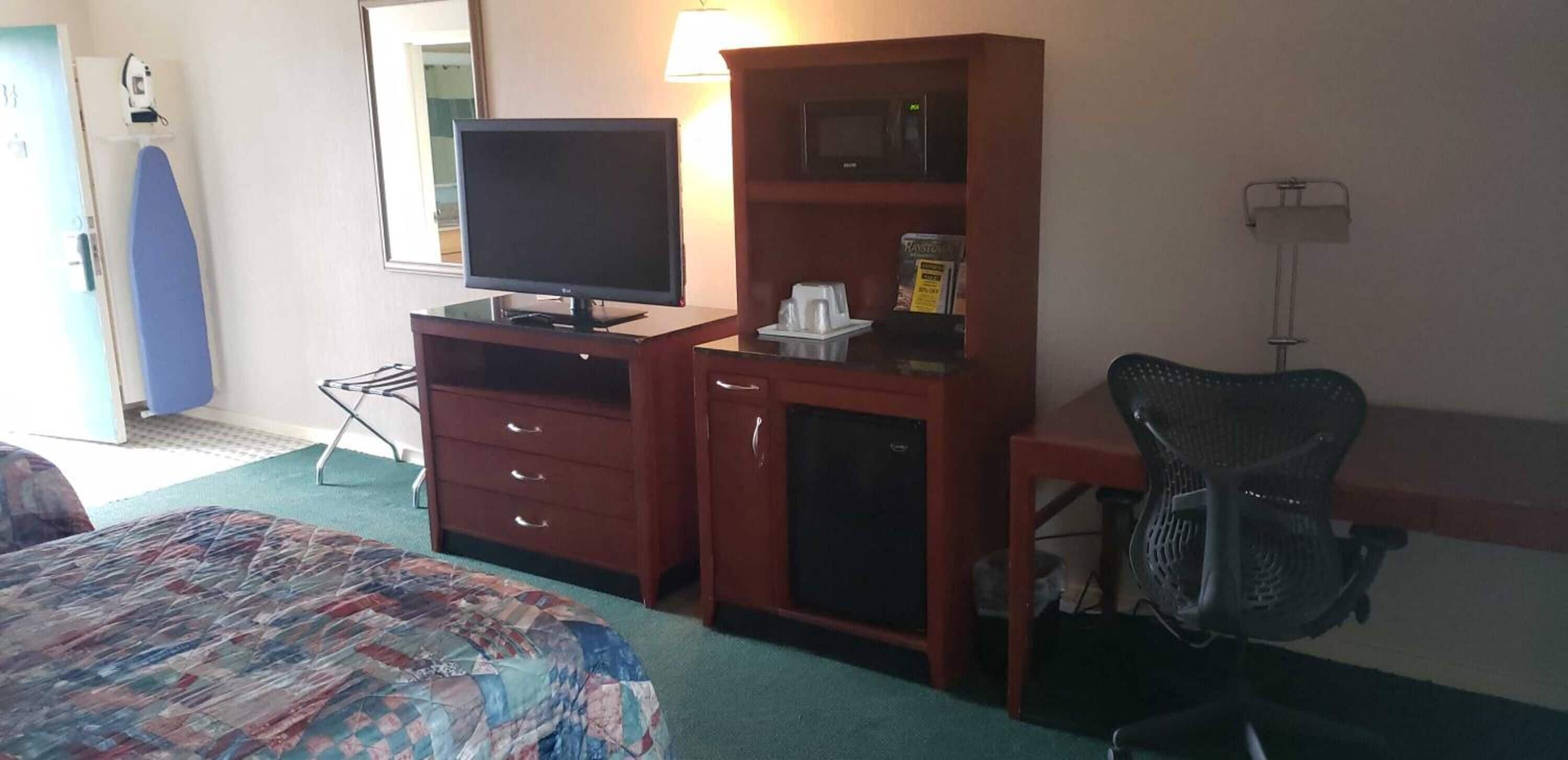 Een hotelkamer met een bed, een bureau met een computer, een stoel, een televisie, een magnetron en een klein kastje.