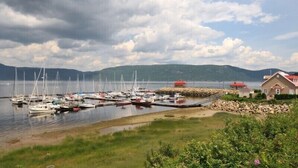 A marina with multiple boats docked, a small building with a red roof, and a rocky shoreline.