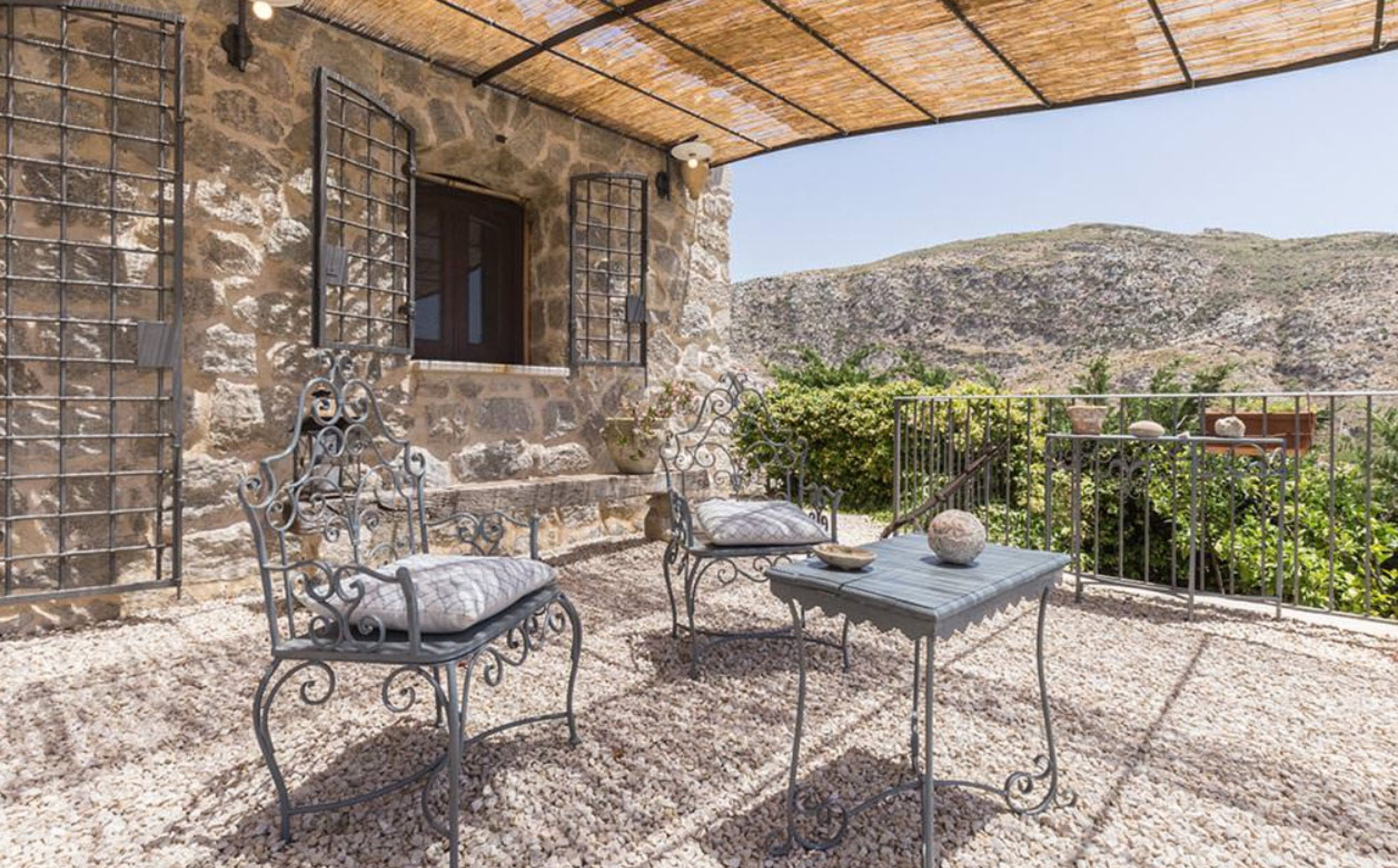 A stone building with a covered patio, two metal chairs, a table, and a view of the hills.