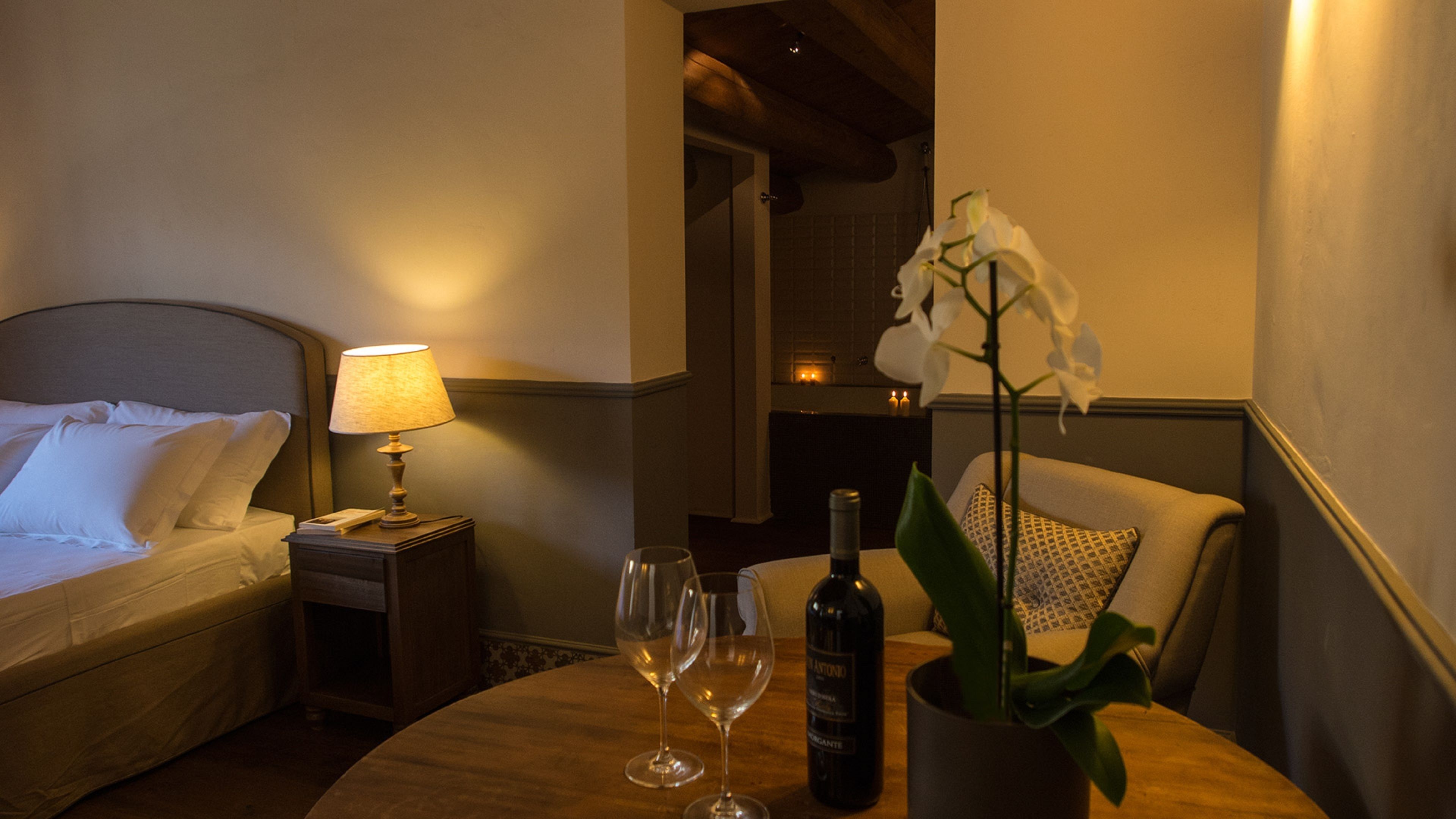A hotel room with a bed, a nightstand with a lamp, two wine glasses, a bottle of wine, and a potted orchid.