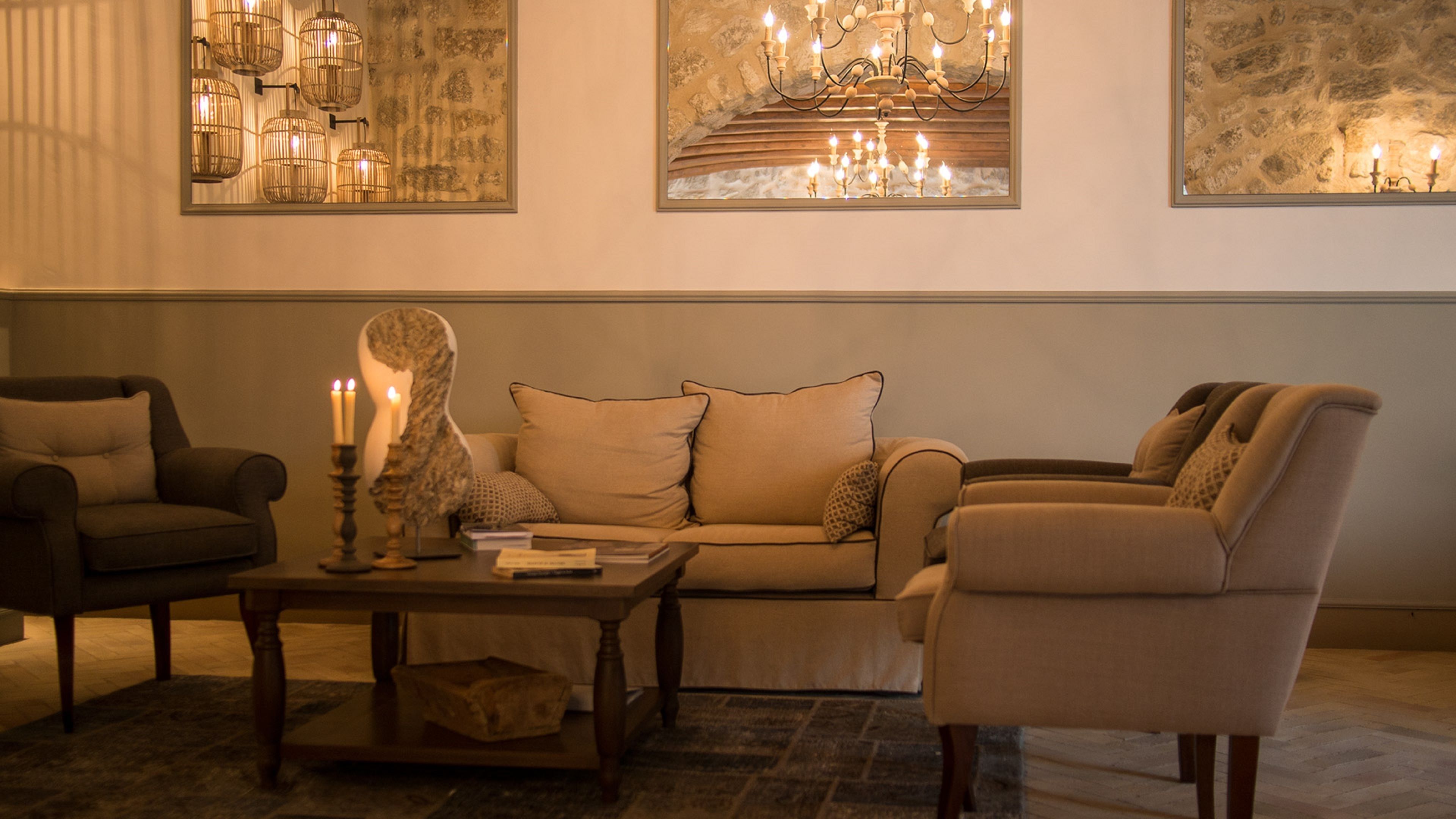 A cozy living room with a sofa, armchairs, and a coffee table. There are framed artworks on the wall and a chandelier.