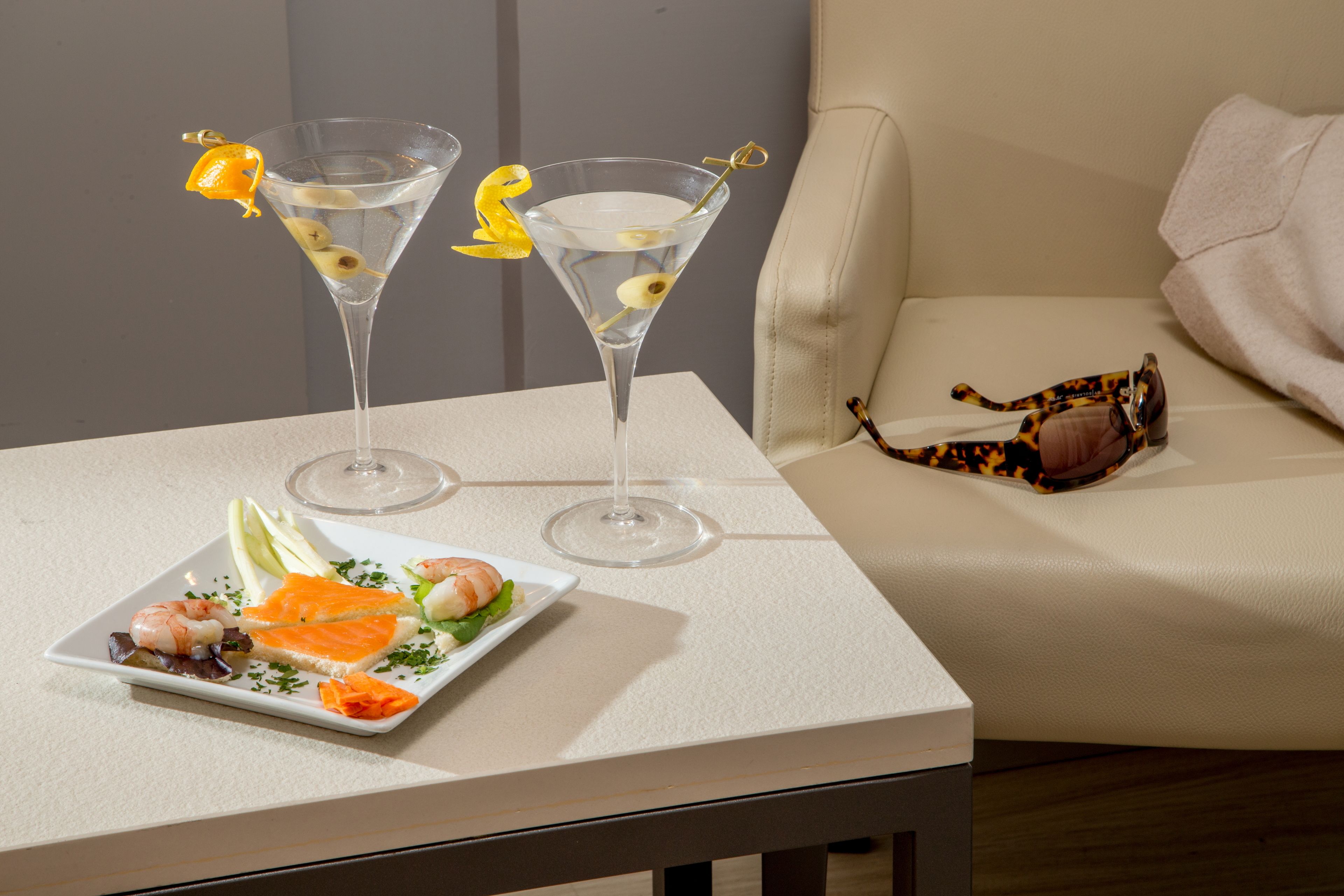 Une table recouverte d’une nappe blanche, avec deux verres à martini, une assiette de nourriture et une paire de lunettes de soleil.