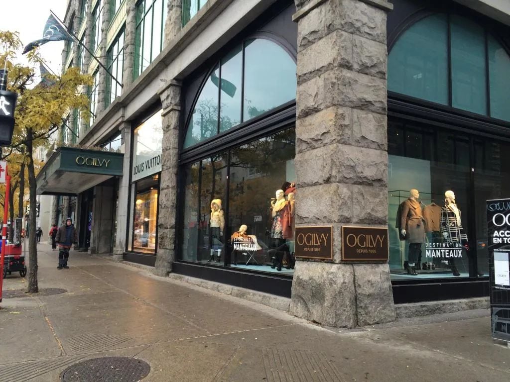 A storefront with large windows displaying mannequins, a sign for 'OGILVY', and a stone pillar.