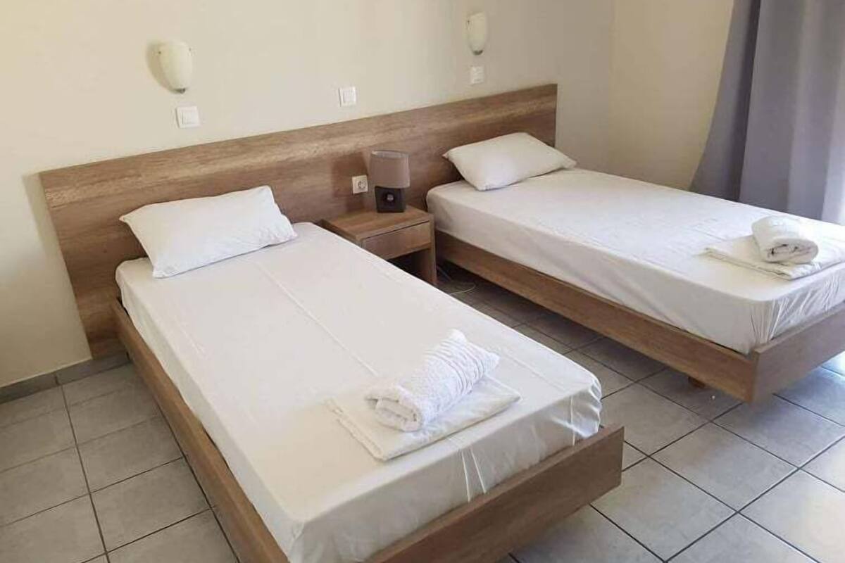 A hotel room with two single beds, each with a wooden headboard and white bedding. There is a nightstand with a lamp between the beds.