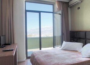 A hotel room with a bed, a desk with a computer and a remote control, and a large window with a view of a city and mountains. - Cajupi (Gjirokastër)