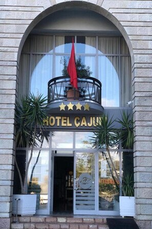 A hotel entrance with a large arched window, a balcony with a red flag, and a sign that reads 'HOTEL CAJUN'. - Cajupi (Gjirokastër)
