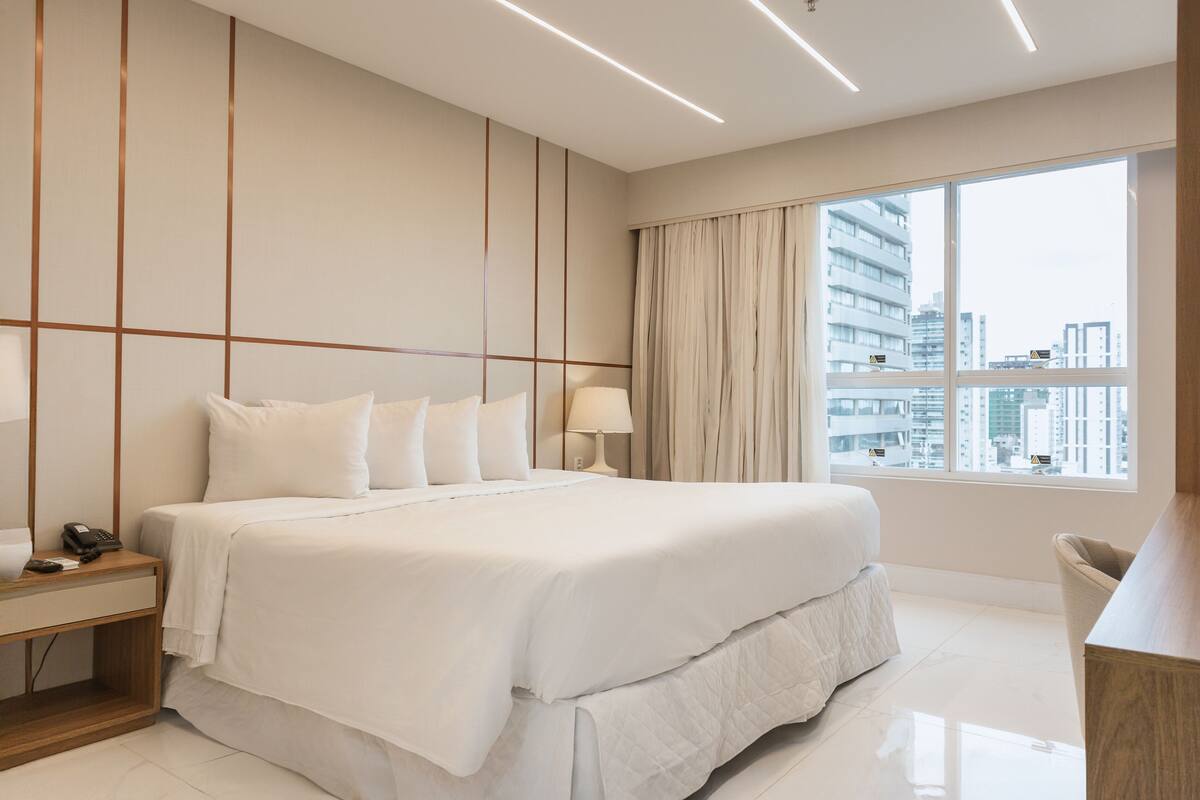 Habitación de hotel moderna con una cama grande, una mesita de noche con lámpara y vistas a la ciudad a través de un ventanal amplio.