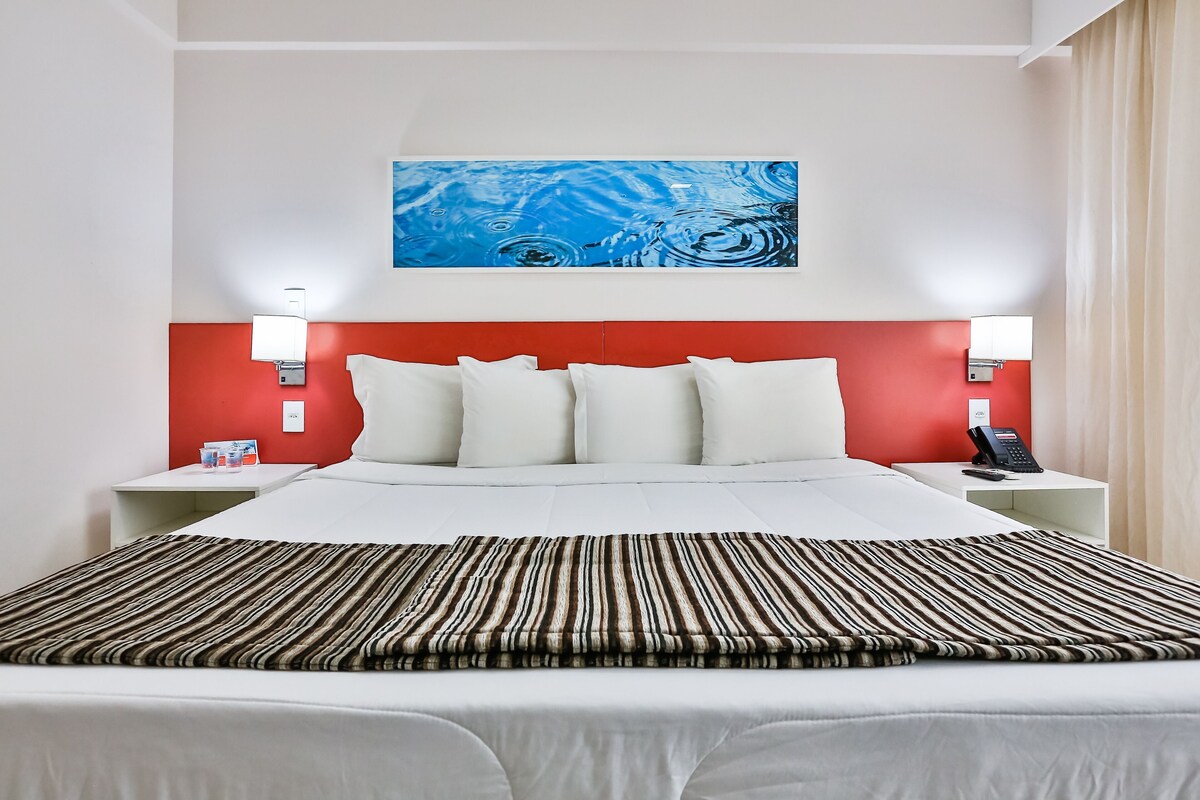 Une chambre d’hôtel avec un lit, du linge de lit blanc, une tête de lit rouge, deux tables de chevet avec des lampes et un tableau abstrait bleu accroché au mur.