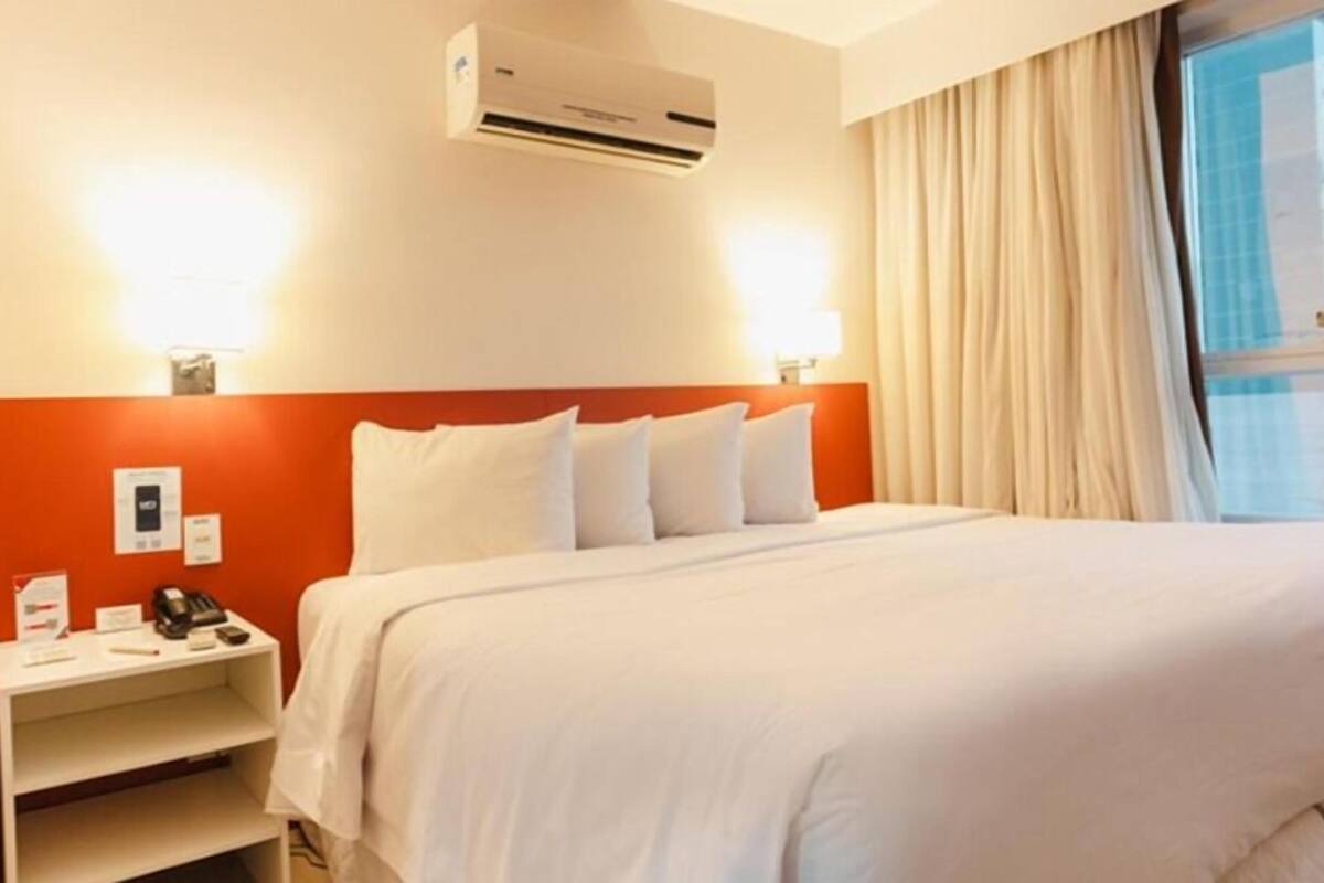 Una habitación de hotel con cama, mesita de noche, aire acondicionado y ventana con cortinas.
