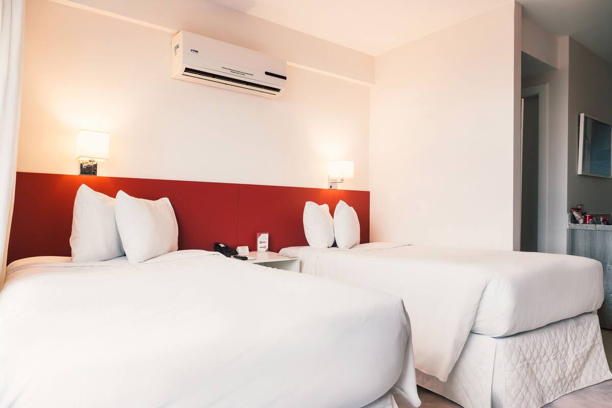 Habitación de hotel con dos camas, cabecera roja, ropa de cama blanca, equipo de aire acondicionado y lámparas de noche.