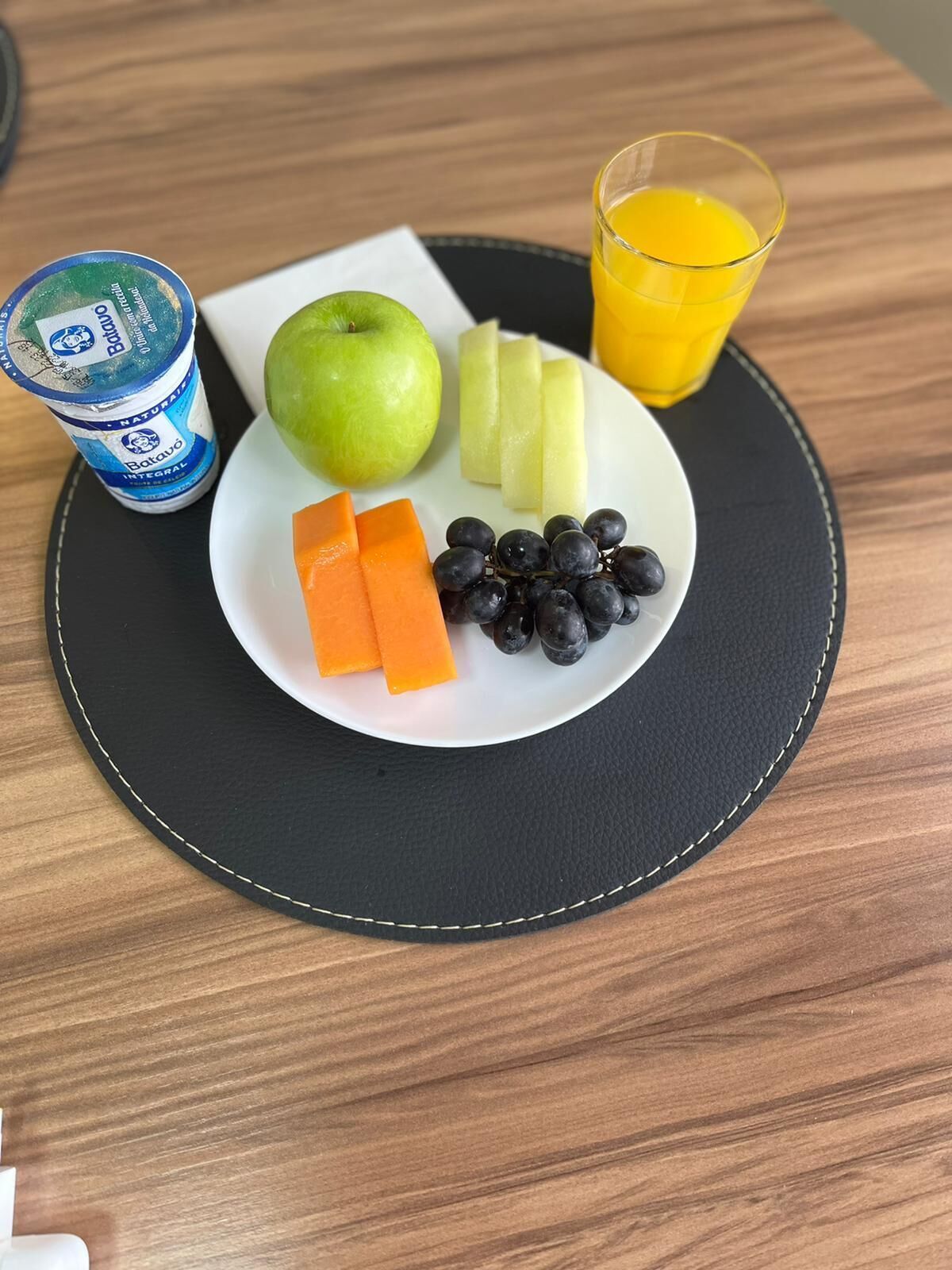 Un plato con una manzana verde, rodajas de fruta amarilla, palitos de zanahoria naranja y arándanos, acompañado de un vaso de zumo de naranja y un recipiente de yogur.