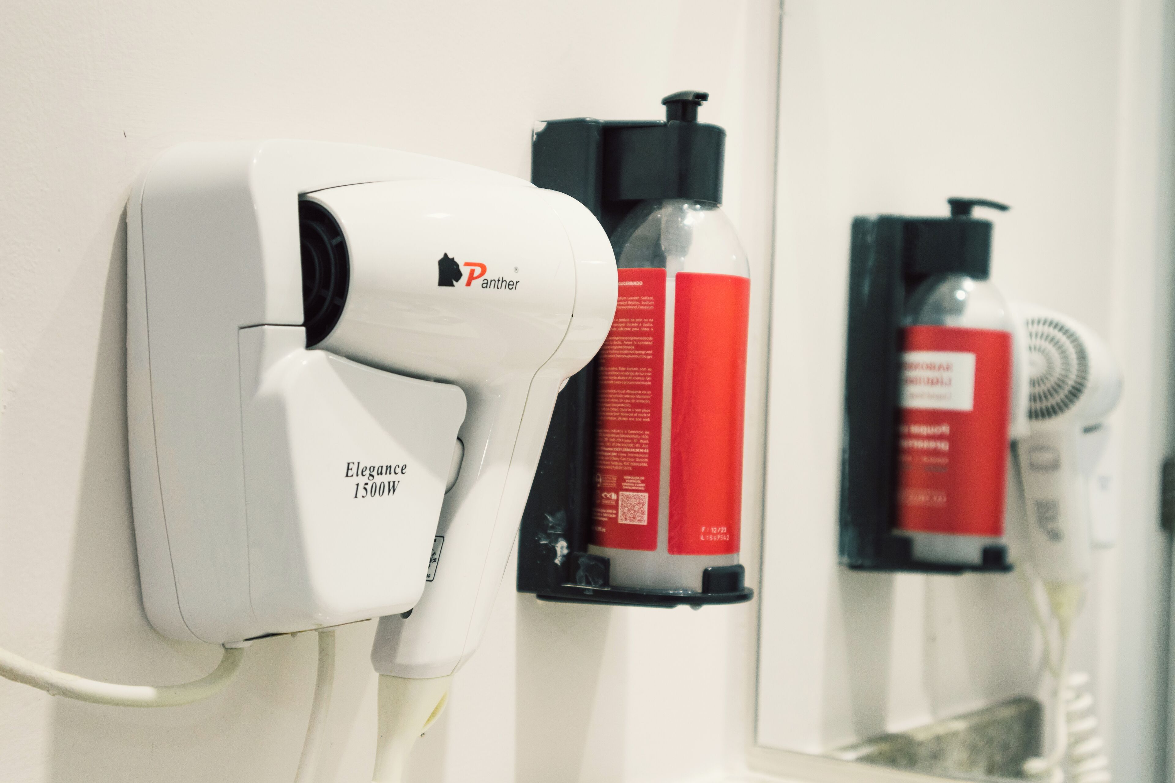A hairdryer mounted on a wall, a red bottle, and a white hairdryer in a bathroom setting.