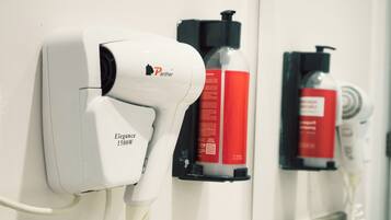 A hairdryer mounted on a wall, a red bottle, and a white hairdryer in a bathroom setting.