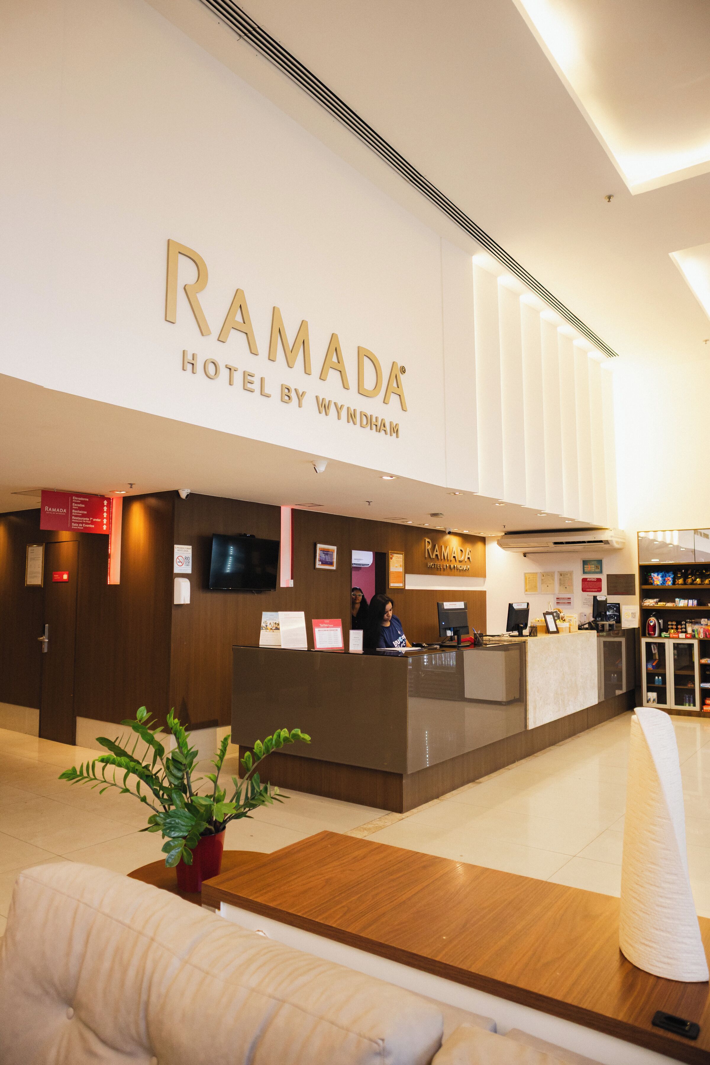 Zona de recepción con mostrador, televisor y un cartel que dice 'RAMADA HOTEL BY WYNDHAM'.