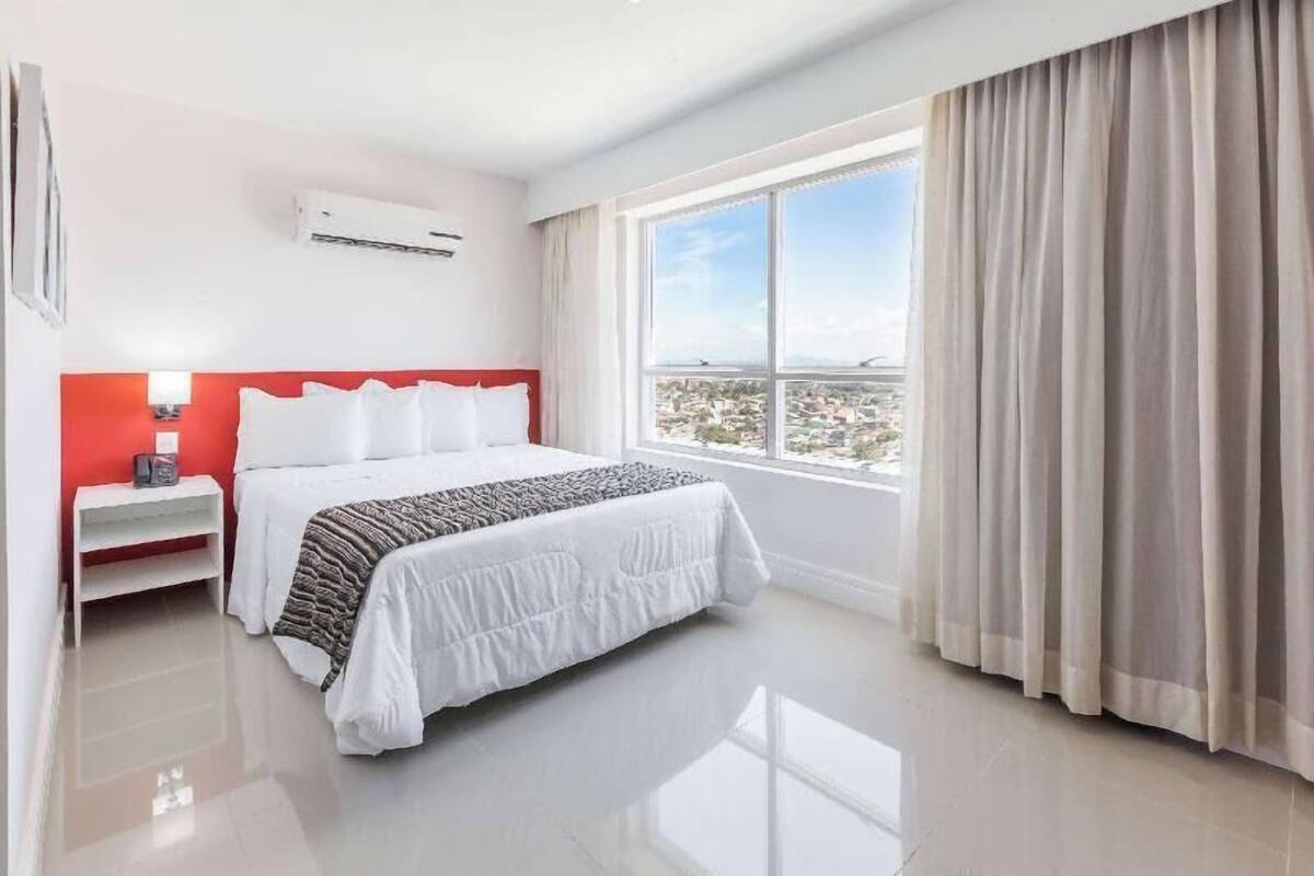 Una habitación de hotel con cama, mesita de noche, ventana con cortinas y aire acondicionado.