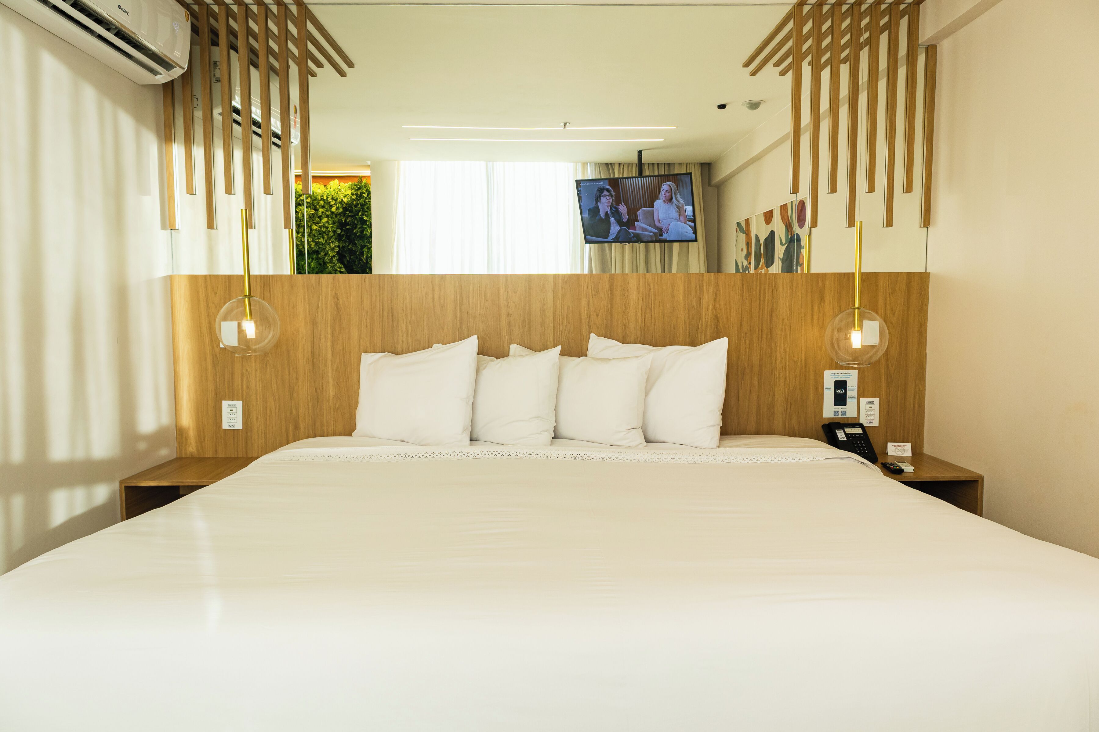A hotel room with a large bed, wooden headboard, two bedside tables with lamps, a TV on the wall, and a window with curtains.