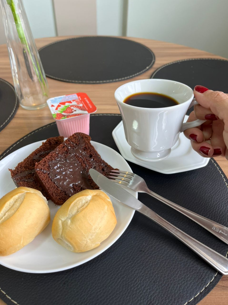 A plate with two chocolate cakes and two buns, a cup of coffee, and a small container of strawberry yogurt on a table.