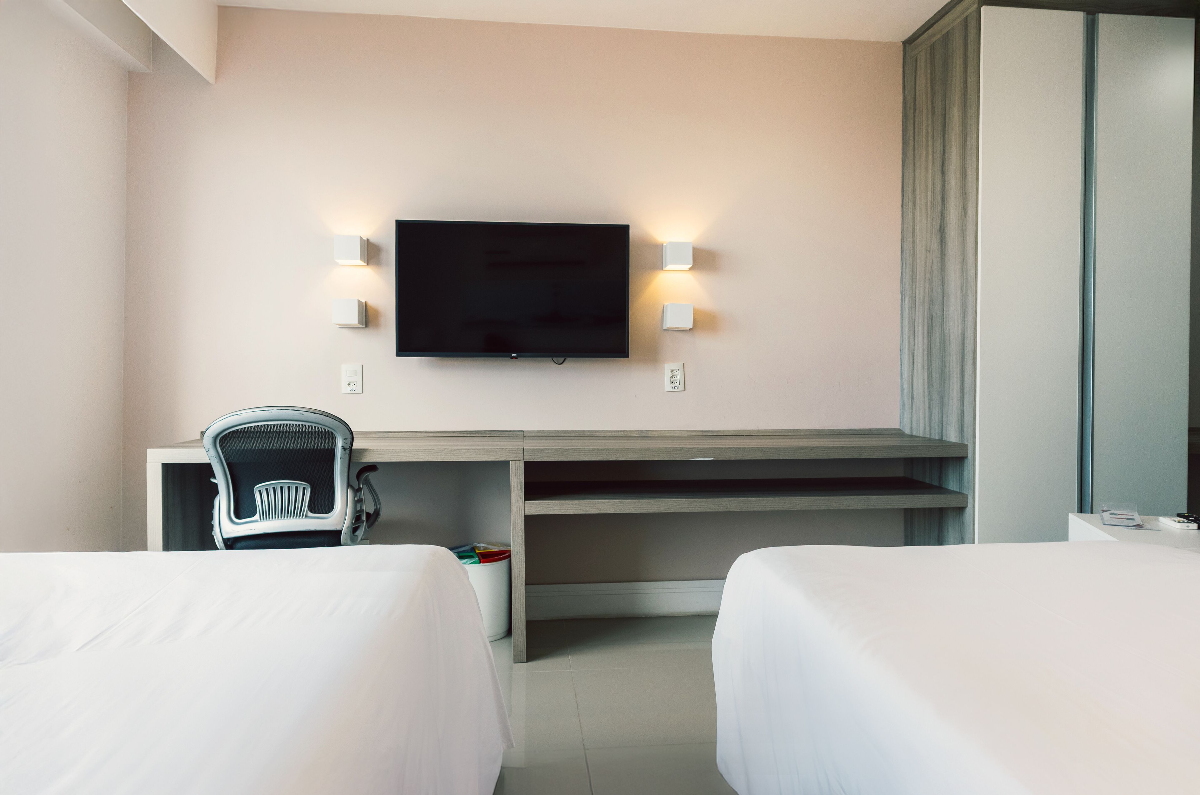 A hotel room with a flat-screen TV mounted on the wall, a desk with a chair, and two beds.