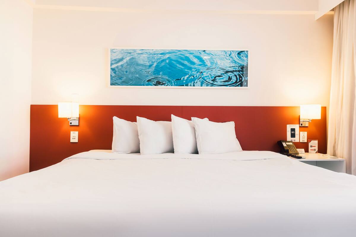 Una habitación de hotel con una cama grande, ropa de cama blanca, cabecera roja y un cuadro abstracto azul en la pared.