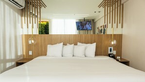 Habitación de hotel moderna con una cama grande, cabecera de madera, dos mesitas de noche con lámparas, un televisor en la pared y una planta pequeña en la esquina.